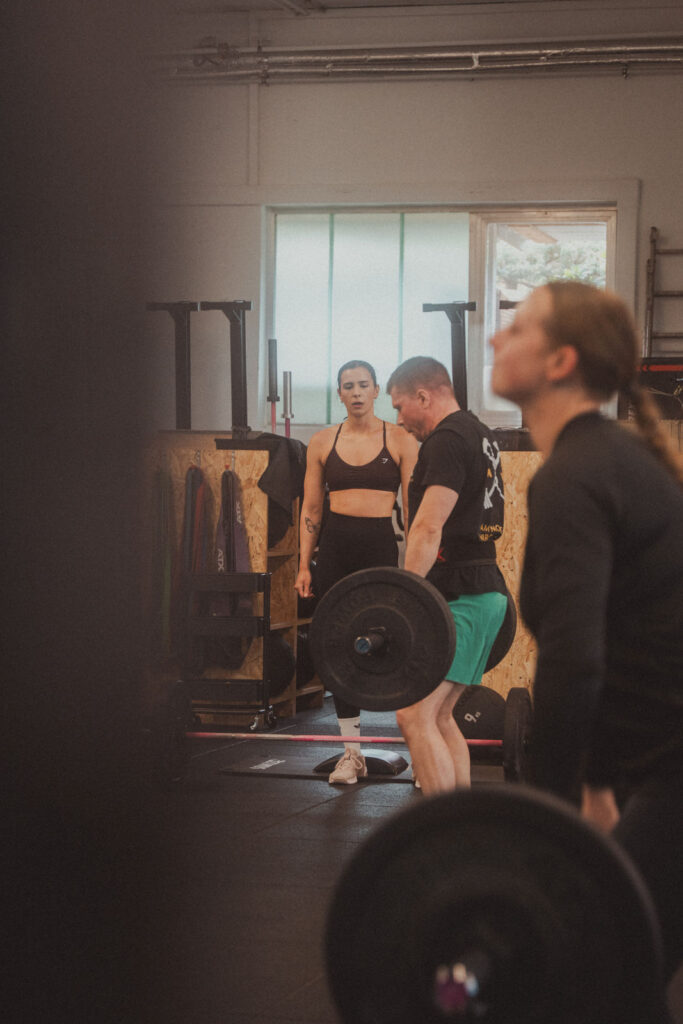 Eine Frau in Sportkleidung steht und beobachtet, wie ein Mann in einem Fitnessstudio eine Langhantel stemmt, und unterstreicht damit die Bedeutung von Kursen und Fertigkeiten. Eine andere Person stemmt Gewichte im Vordergrund, während im Hintergrund Fitnessgeräte und natürliches Licht zu sehen sind.