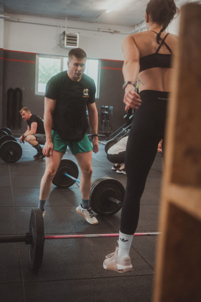 Zwei Personen trainieren in einem Fitnessstudio mit Hanteln auf dem Boden und zeigen ihre Kurse & Fähigkeiten; eine steht, während die andere, in einem Sport-BH und Leggings, sich auf das Heben oder Springen vorbereitet. Im Hintergrund sind andere Fitnessstudio-Besucher zu sehen.