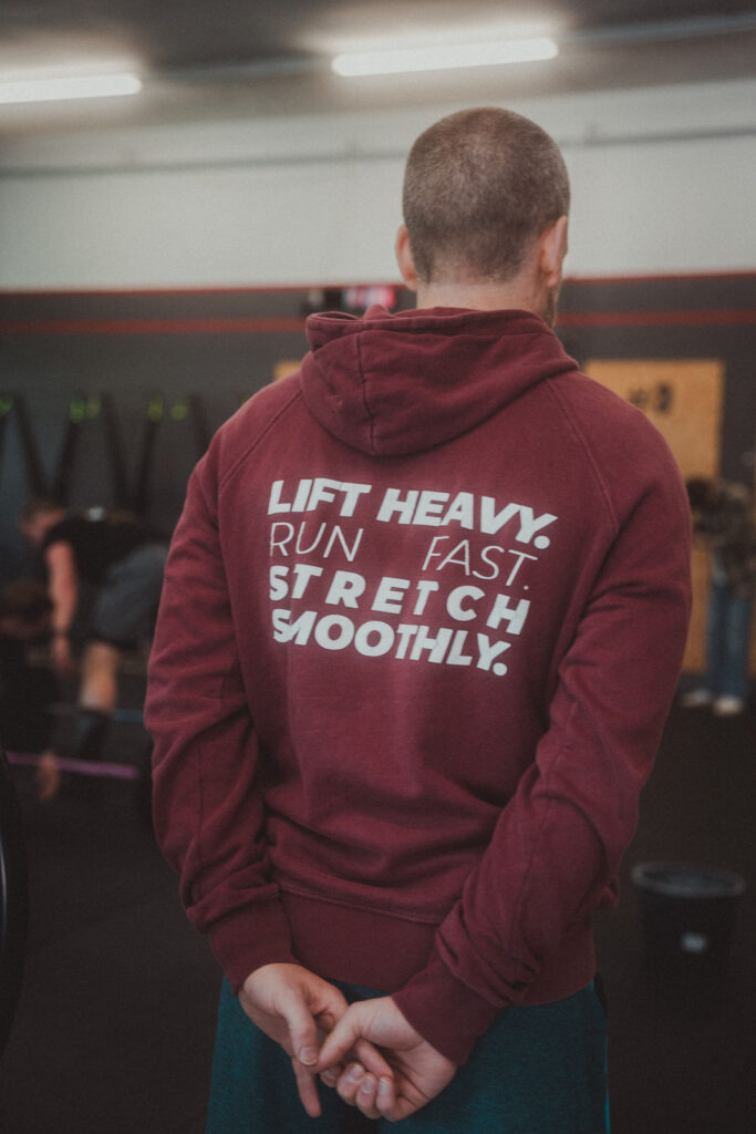Eine Person in einem kastanienbraunen Kapuzenpullover mit der Aufschrift LIFT HEAVY. SCHNELL LAUFEN. STRETCH SMOOTHLY. steht in einem Fitnessstudio, die Hände hinter dem Rücken verschränkt, während andere in der Nähe trainieren - perfekt, um neue Fitness-Angebote zu präsentieren.