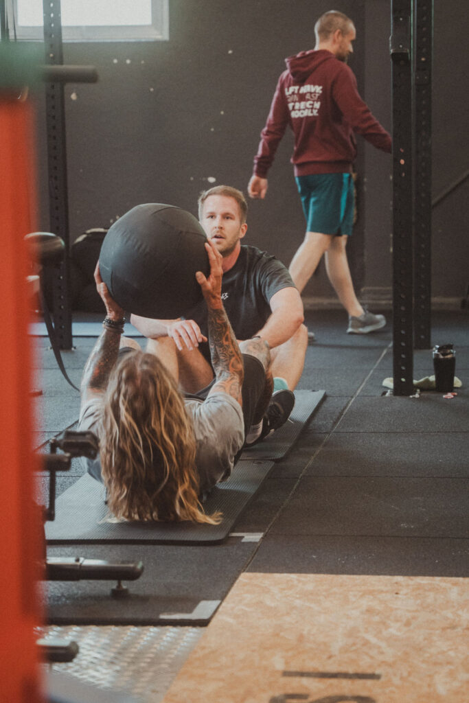 Eine Person mit langen Haaren und Tattoos macht Sit-ups und hält einen Medizinball, während eine andere Person ihre Füße stützt. Im Hintergrund geht ein Mann in einem roten Kapuzenpulli an Fitnessgeräten vorbei und sieht sich die neuesten Angebote an.