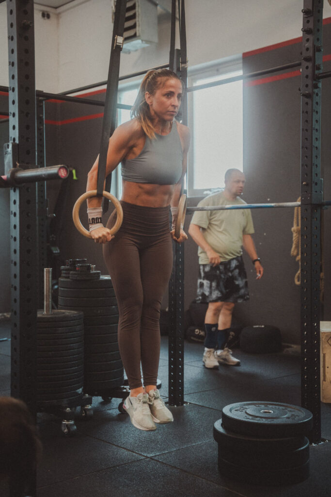 Eine Frau in Sportkleidung führt in einem Fitnessstudio eine Ringdip-Übung an Gymnastikringen aus. Konzentriert und stark trainiert sie zwischen verschiedenen Angeboten für Fitnessbegeisterte. Im Hintergrund sind Fitnessgeräte und eine weitere Person zu sehen.