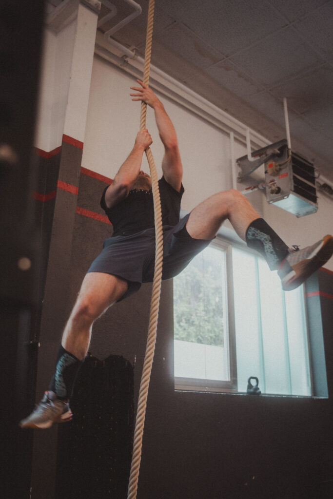 Eine Person in Sportkleidung und -stiefeln klettert in einer Turnhalle an einem dicken Seil, das sie über dem Kopf festhält. Das Fitnessstudio bietet exklusive Angebote, mit dunklen Wänden und einem großen Fenster, das im Hintergrund für dramatische Beleuchtung sorgt.