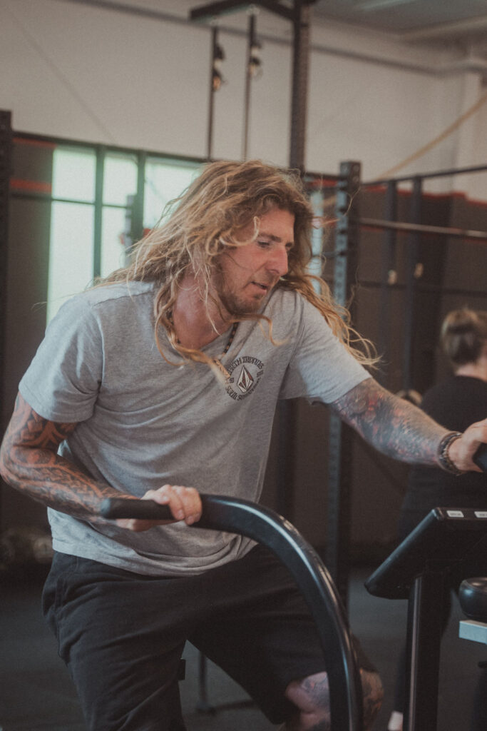 Eine Person mit langem, gewelltem blondem Haar und tätowierten Armen trainiert auf einem stationären Fahrrad in einem Fitnessstudio und stellt ihre Fitnessfähigkeiten unter Beweis. Sie trägt ein graues T-Shirt und schwarze Shorts und fährt zwischen Fitnessgeräten und einer anderen Person, die an Gruppenkursen teilnimmt.