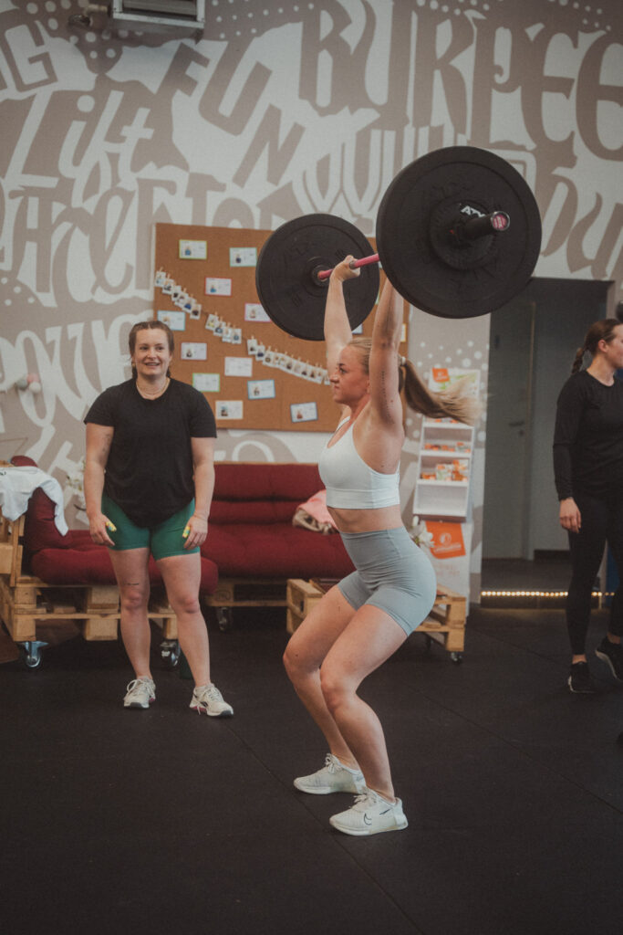 Eine Frau in Sportkleidung hebt in einem Fitnessstudio eine Langhantel über den Kopf, während eine andere Frau in Shorts und schwarzem Hemd lächelt und zusieht. Motivierende Worte schmücken die Wand, ein rotes Sofa steht in der Nähe, und für Mitglieder gibt es exklusive Angebote.