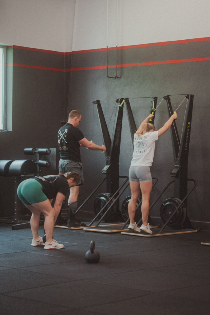 Zwei Personen arbeiten an ihren Skierg-Fähigkeiten, während eine andere sich in einem Fitnessraum mit schwarzen und roten Wänden neben einer Kettlebell beugt. Sonnenlicht fällt ein und erhellt einen Teil des Raums - ein energiegeladener Ort für Kurse & Skills.