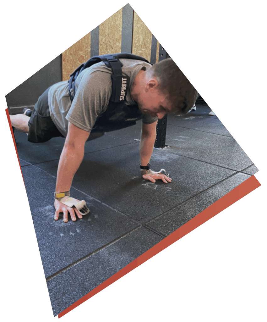 Eine Person, die eine Gewichtsweste und einen Handschuh trägt, macht einen Liegestütz auf dem Boden eines Fitnessstudios, wobei Kreidemarkierungen auf der Matte zu sehen sind, die Kurse & Fertigkeiten zeigen. Das Bild ist diagonal gedreht.