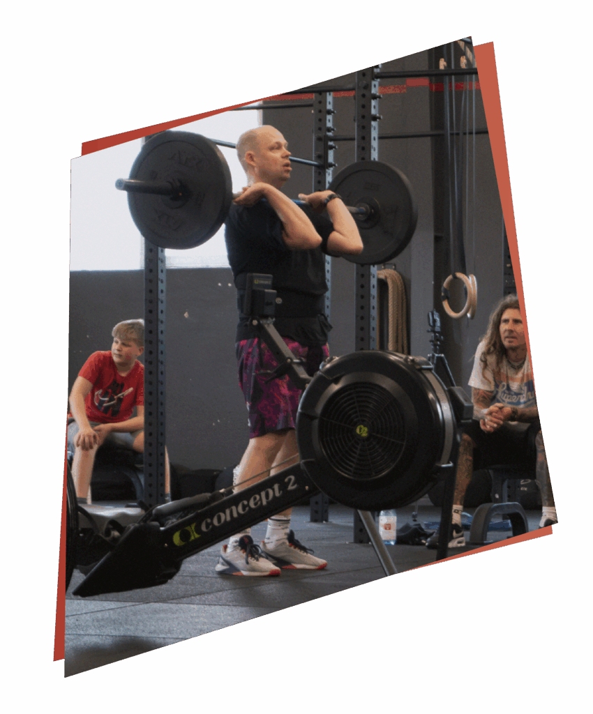 Eine Person hebt eine Langhantel in einem Fitnessstudio, während andere zuschauen und ihre Kurse & Fertigkeiten präsentieren. Im Vordergrund steht ein Concept2 Rudergerät, im Hintergrund sind verschiedene Fitnessgeräte zu sehen.