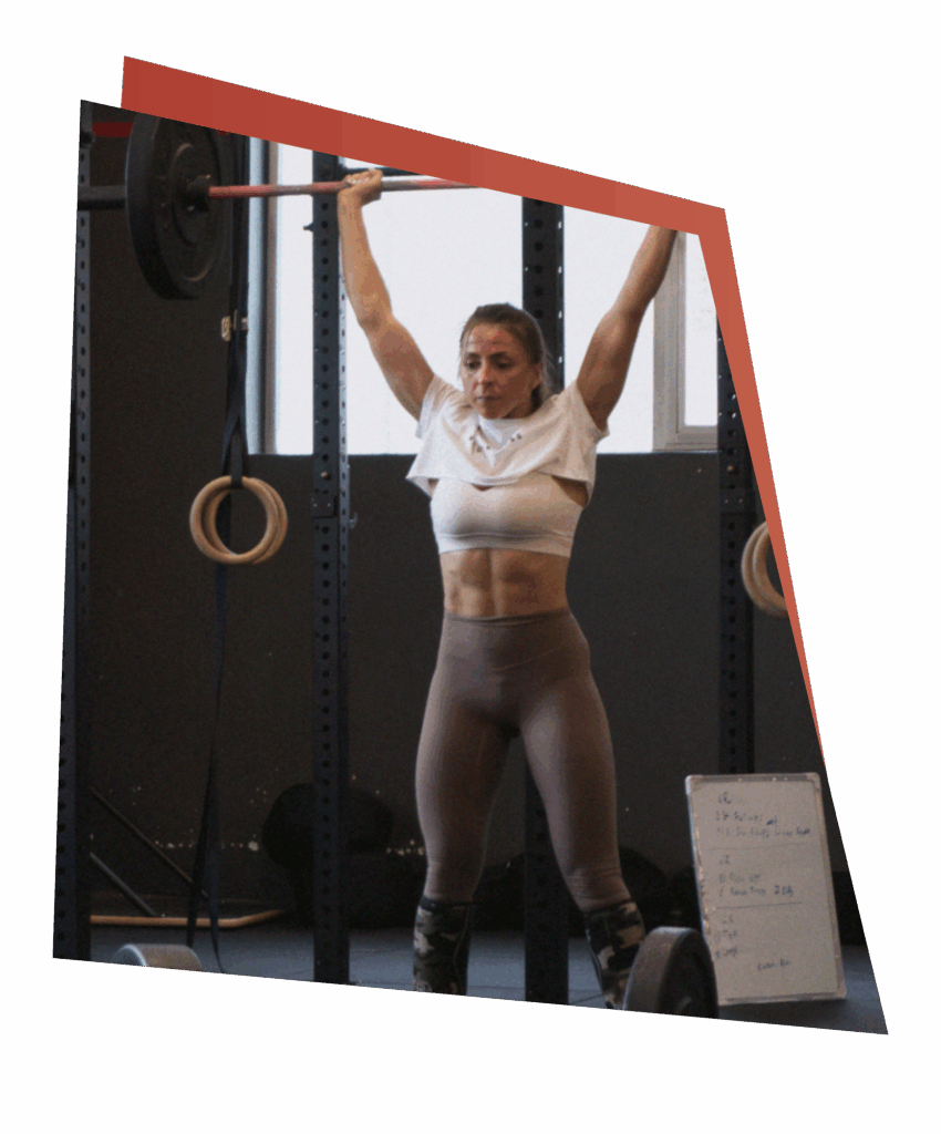 Eine Frau in Trainingskleidung hebt in einem Fitnessstudio eine Langhantel über den Kopf und demonstriert damit ihre Kraft und ihr Können. Sie steht zwischen Gymnastikringen, in der Nähe befinden sich eine Tafel und Gewichte - eine inspirierende Szene für die Entwicklung von Kursen und Fertigkeiten. Das Bild hat einen schrägen Rand.