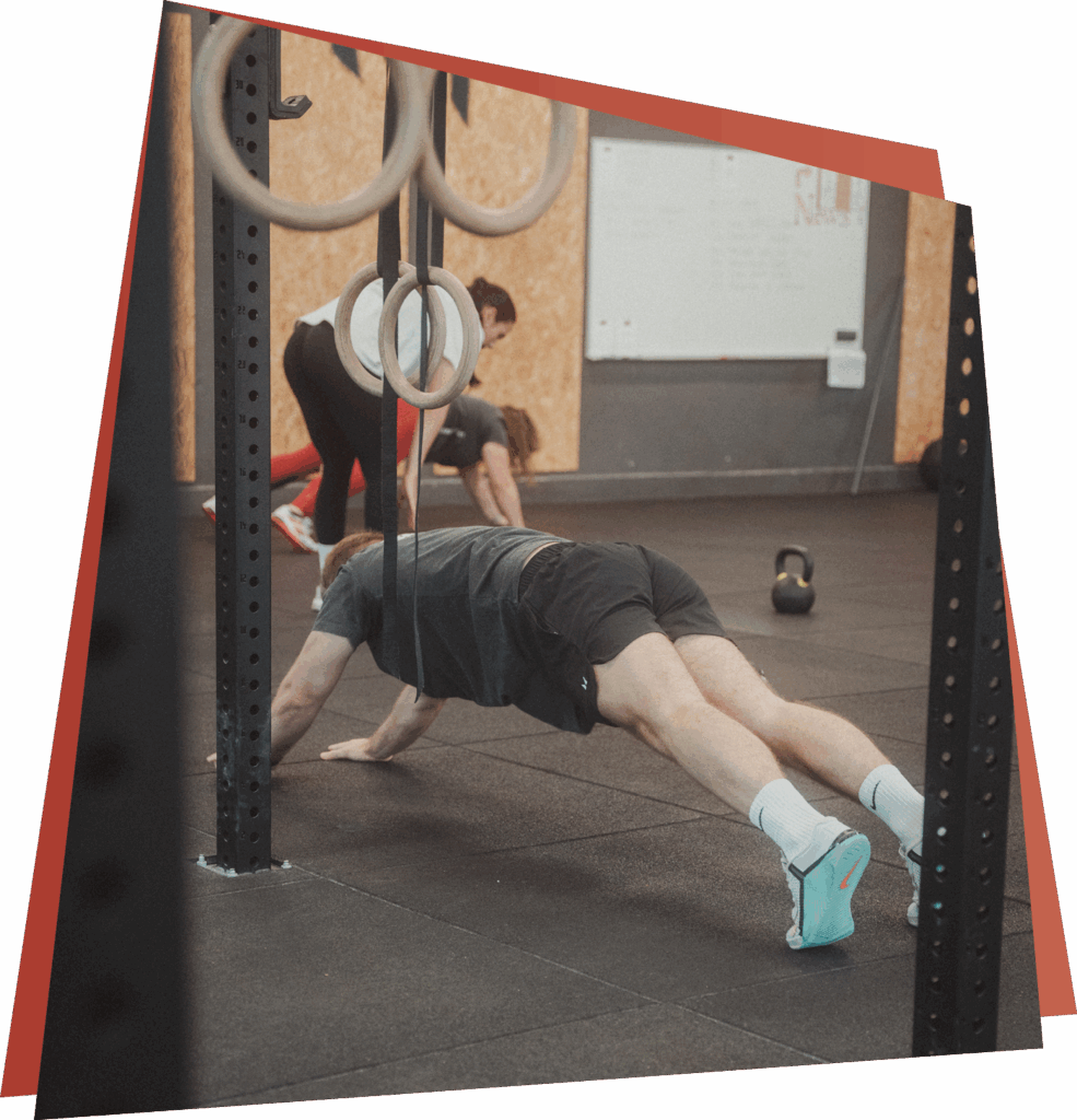 Eine Person in Sportkleidung macht Liegestütze auf einem Turnhallenboden in der Nähe von Gymnastikringen und zeigt ihre Fähigkeiten, während eine andere Person sich dehnt und eine Kettlebell im Hintergrund steht - perfekt für alle, die anspruchsvolle Kurse und Fähigkeiten suchen.