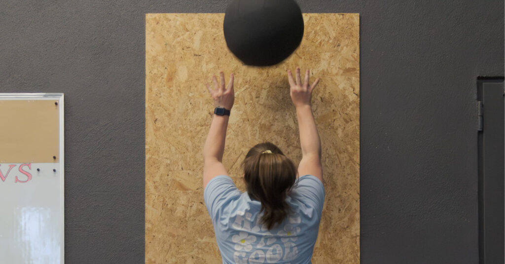 Eine Person in einem hellblauen Hemd wirft einen schwarzen Medizinball gegen ein Holzbrett, das an einer dunklen Wand in einem Fitnessstudio angebracht ist, und zeigt dabei ihre Kurse und Fähigkeiten.