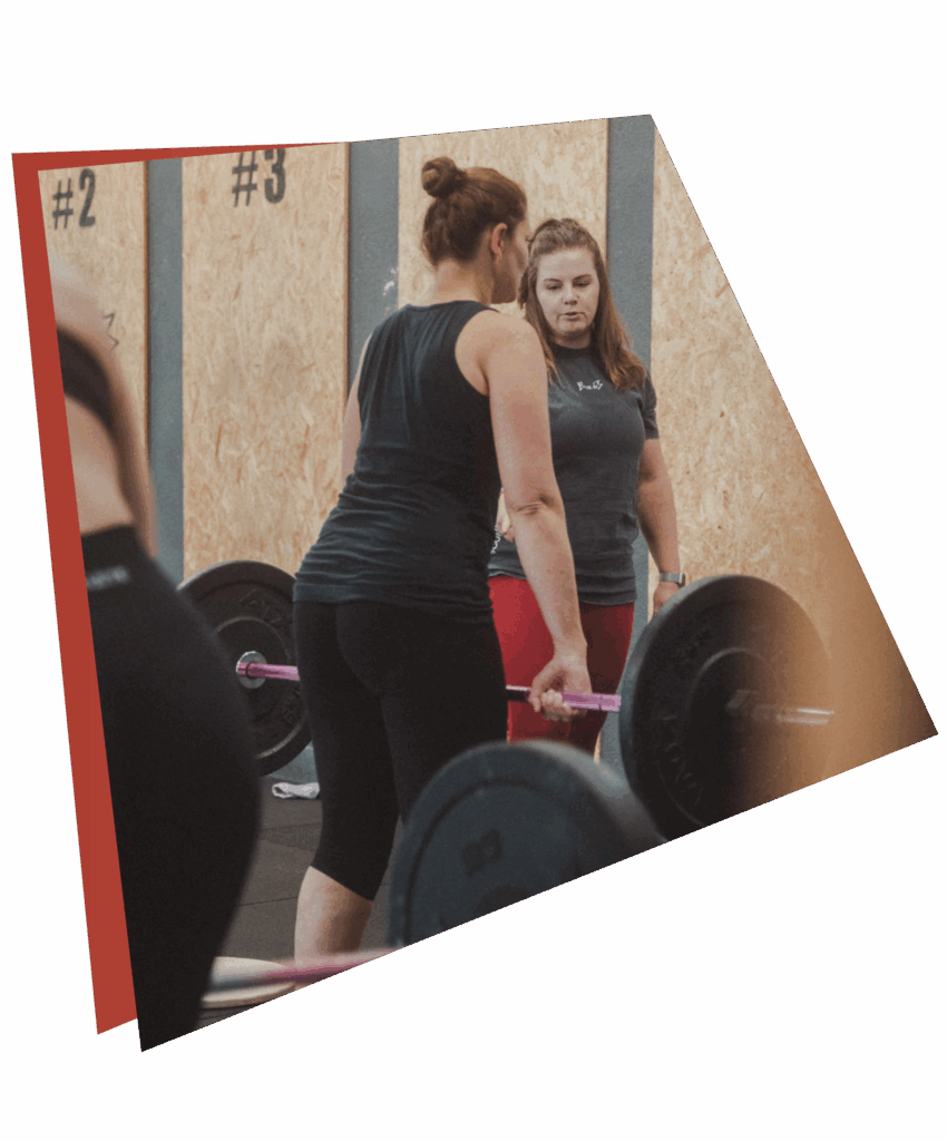 Zwei Frauen in Sportkleidung befinden sich in einem Fitnessstudio. Die eine hebt eine Langhantel, während die andere sie beobachtet und anleitet - dies unterstreicht die Entwicklung von Kursen und Fähigkeiten. Im Hintergrund sind hölzerne Wandtafeln mit nummerierten Abschnitten zu sehen.