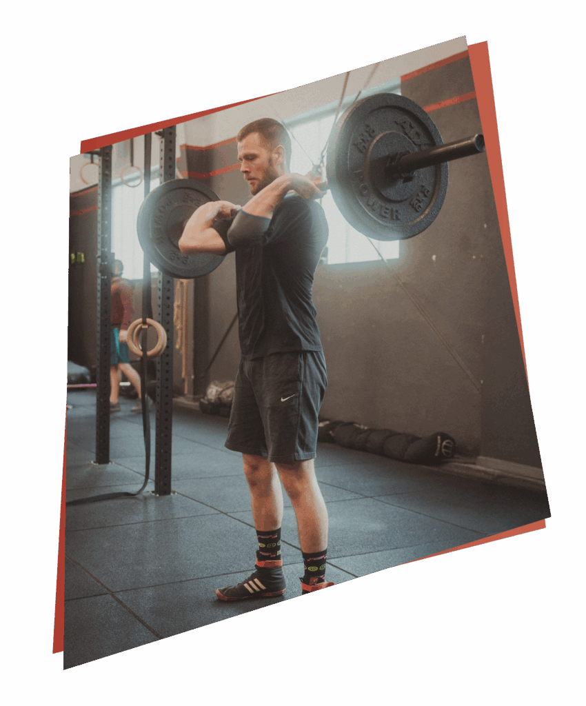 Ein Mann in Sportkleidung führt eine Kniebeuge mit einer belasteten Langhantel in einem Fitnessstudio aus, wobei er sowohl Kurse als auch Fertigkeiten zeigt. Er steht auf einem Gummiboden in der Nähe von Gymnastikringen und anderen Geräten.