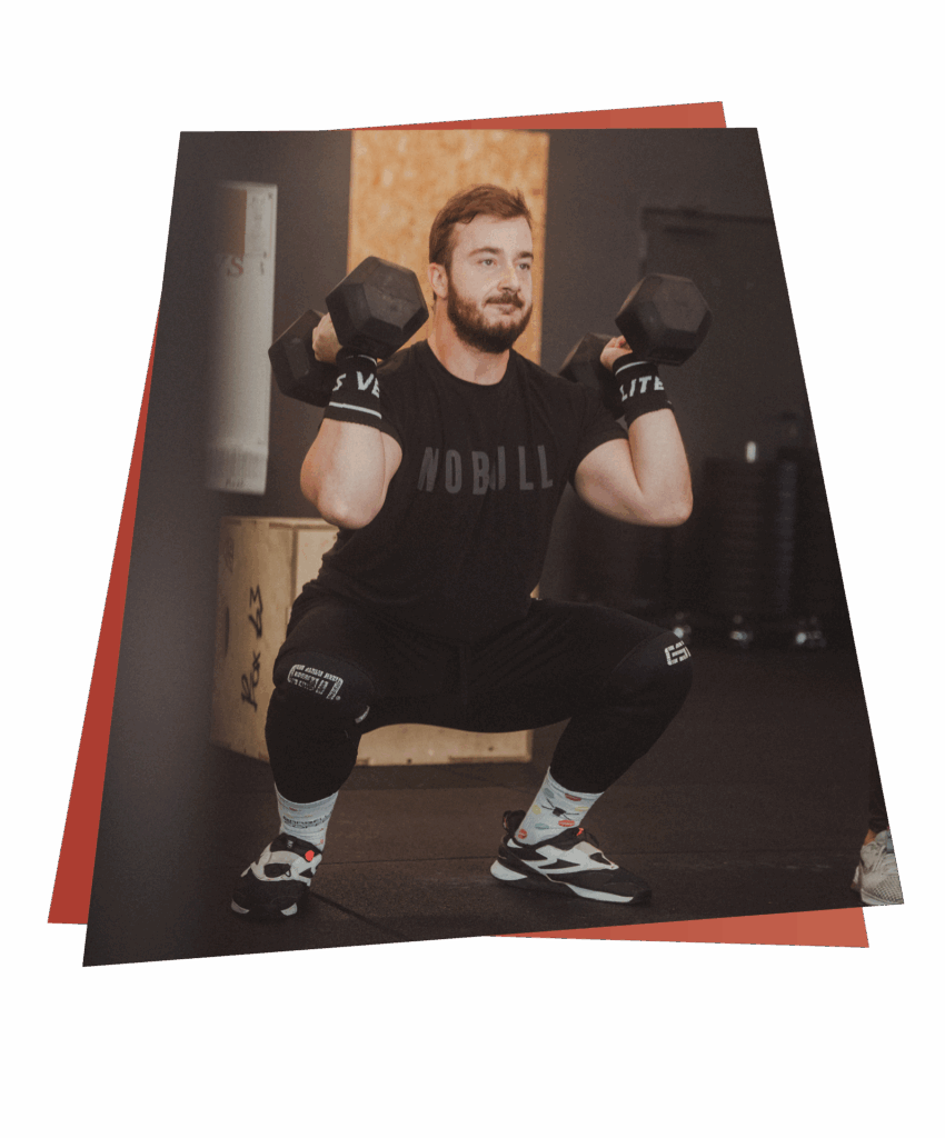 Ein Mann mit Bart geht in einem Fitnessstudio in die Hocke, während er in jeder Hand eine Kurzhantel auf Schulterhöhe hält und seine Kurse und Fähigkeiten präsentiert. Er trägt ein schwarzes NOBULL-Shirt, eine schwarze Hose und Sportschuhe, umgeben von Fitnessgeräten.