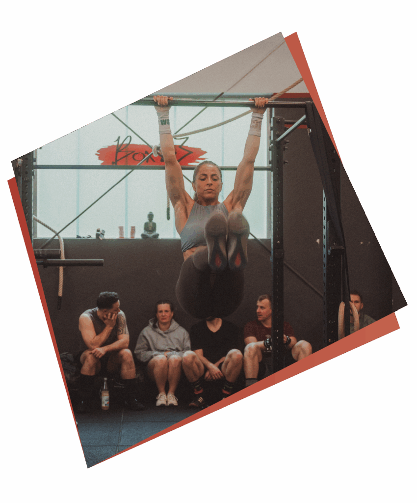 Eine Frau demonstriert Kurse & Fertigkeiten, indem sie einen hängenden Beinheber an einer Klimmzugstange in einem Fitnessstudio ausführt, während vier Personen auf einer Bank im Hintergrund sitzen und ihr beim Training zusehen.